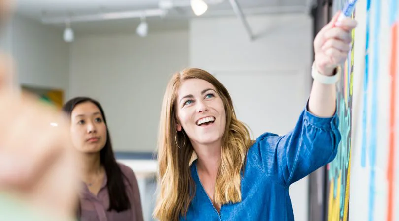 Two colleagues at a whiteboard covered in colored sticky notes; the woman in front, in a denim shirt, is smiling and reaching up to write.