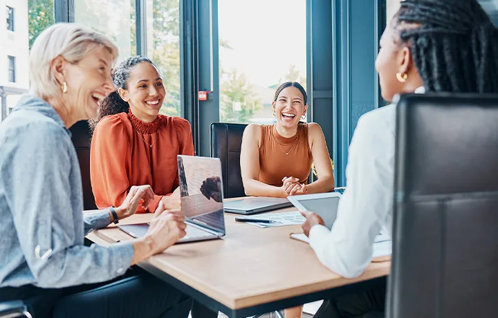 Four colleagues laughing together around a conference table with laptops, in front of floor-to-ceiling windows.