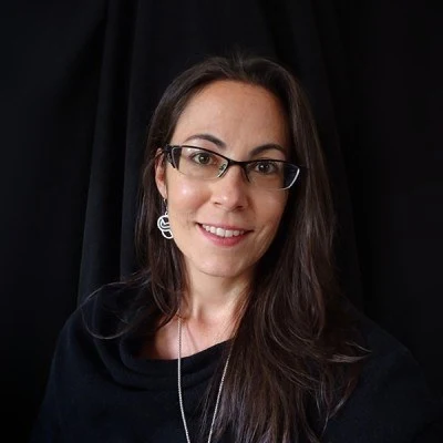 Brianne Hans, smiling, with long dark hair and glasses, in black against a dark backdrop.