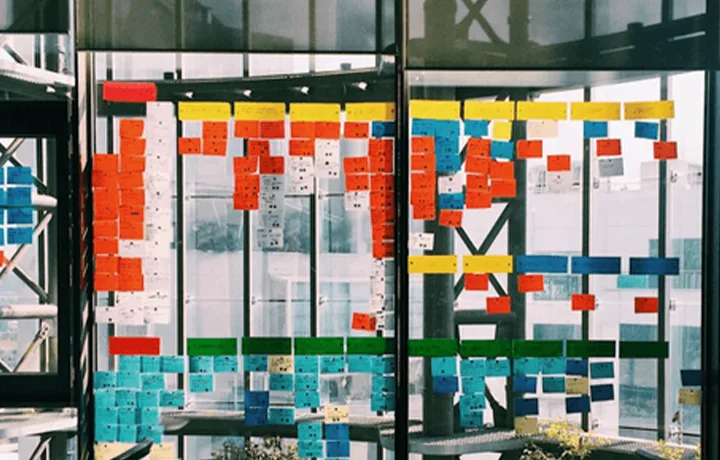A glass wall covered in a grid of colored sticky notes — red, orange, yellow, blue and green rows — used for planning.