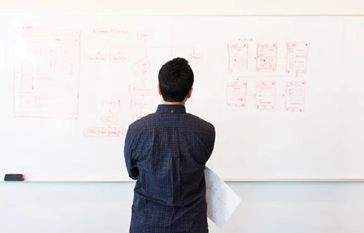 A person seen from behind, studying a whiteboard covered in hand-drawn website wireframes and flow diagrams in red marker.