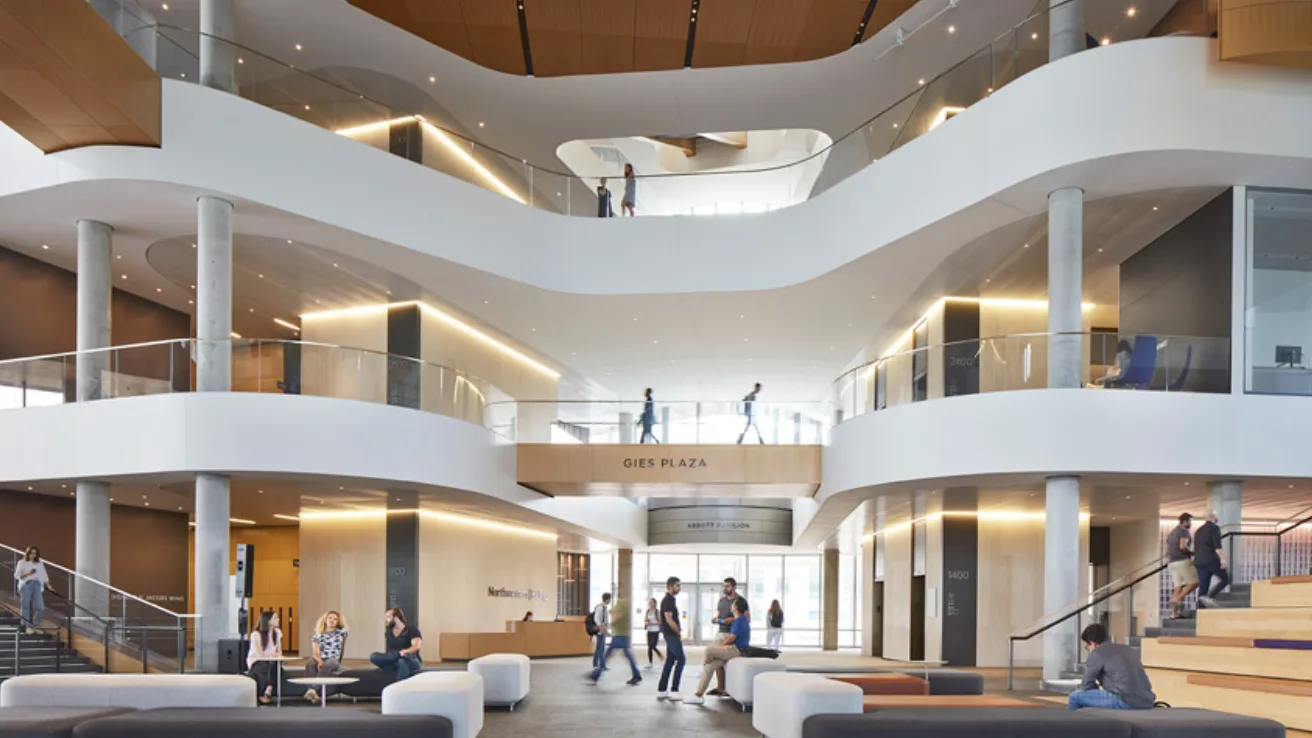 The atrium of Kellogg's Global Hub — curving white balconies stacked over Gies Plaza, with students crossing a bridge and gathered on low seating below.