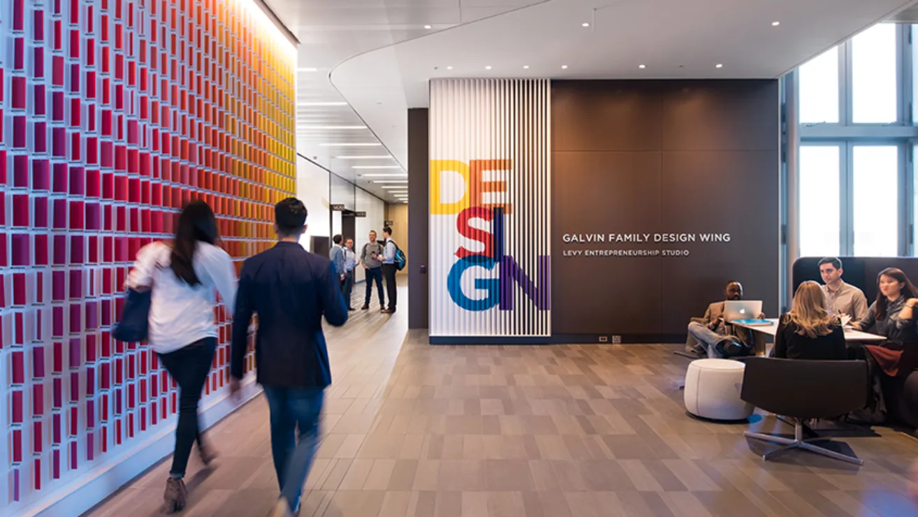 The Galvin Family Design Wing inside Kellogg's Global Hub — a wall of red-to-orange tiles on the left, a striped DESIGN supergraphic on the far wall beside the wing's signage, and students walking and working at a table.