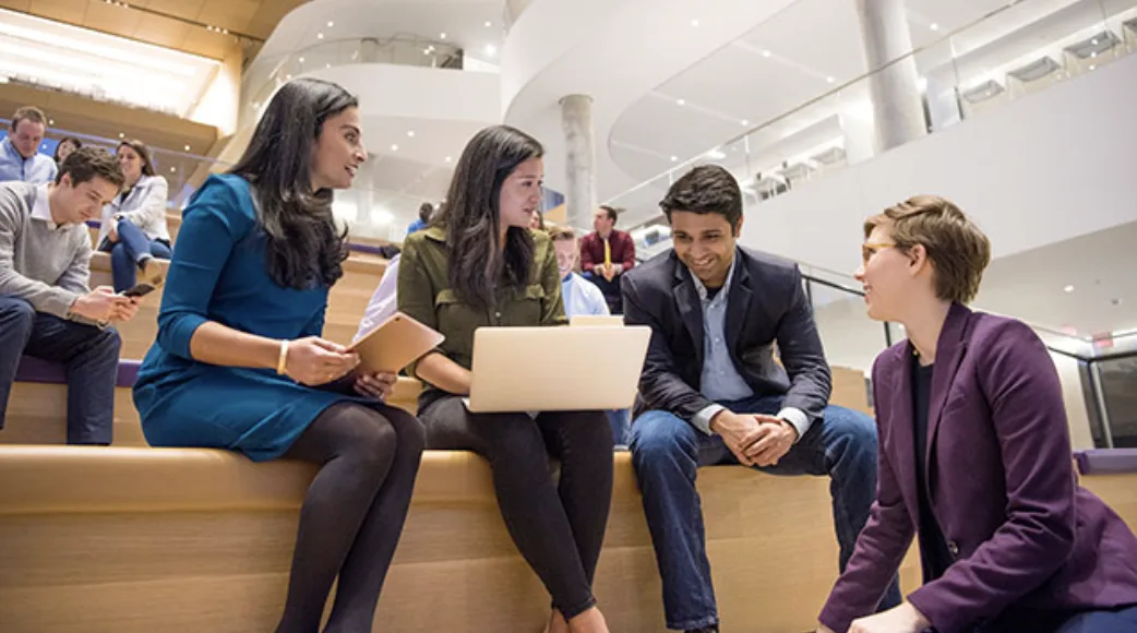 Four students in conversation on the tiered wooden seating inside Kellogg's Global Hub, one with a laptop open.